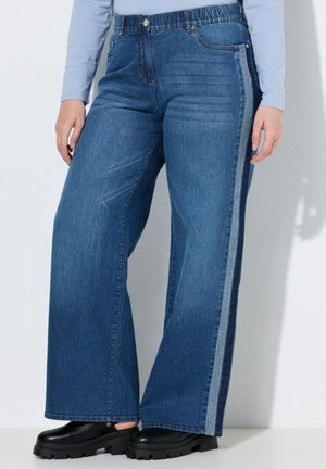 Person iført blå viddebenede jeans med lyse og mørke denim sidelister og sorte sko med chunky såler, står op ad en hvid væg.