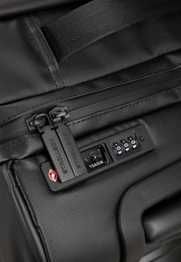 Angolo di una valigia nera con doppi cursori della zip con il marchio "Strellson", una serratura TSA e una texture a motivi sulla superficie.