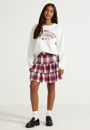 Witte sweatshirt met "Fijne Feesten" print in rood en zwart; gelaagde rood-witte geruite rok; zwarte instappers; witte sokken.