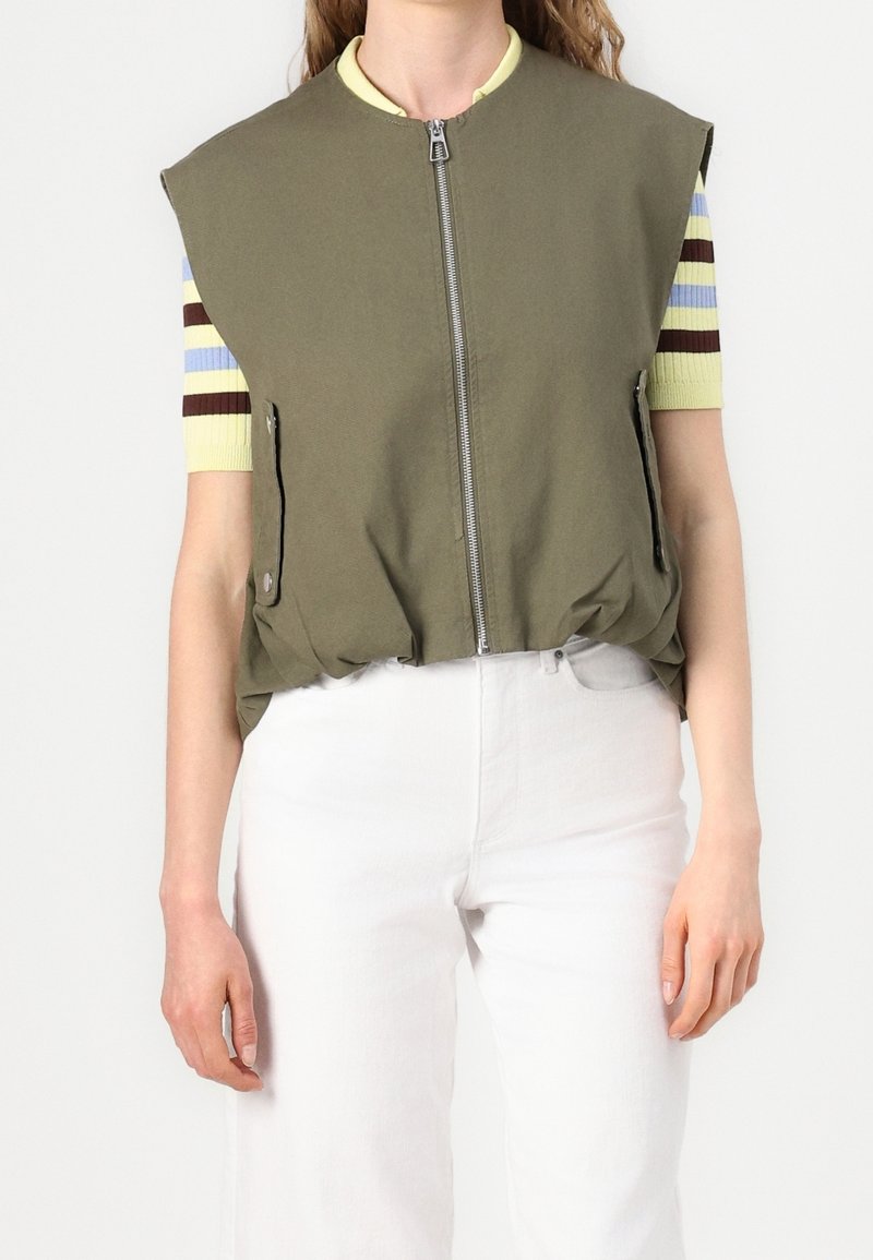 Olivgrüne Weste mit hohem Kragen, Frontreißverschluss und Seitentaschen; getragen über einem gestreiften Kurzarmshirt und weißen Hosen.