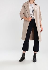 Beige pláštěnka s velkým límcem, tříčtvrtečními rukávy a otevřeným předním dílem, doplněná tmavými rozšířenými kalhotami a černými kotníkovými botami.