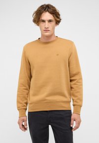 Musztardowa bluza wykonana z teksturowanej tkaniny, z okrągłym dekoltem i ściągaczami na rękawach, noszona na ciemnych spodniach. Subtelny detal z logo na piersi.