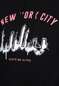 Černá mikina s kapucí a grafikou bílé siluety newyorské panorama, růžový text "NEW YORK CITY" nahoře a "KEEPS ME ALIVE" pod siluetou.