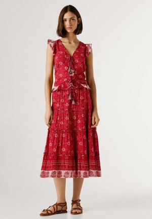 Femme portant une robe rouge à fleurs sans manches avec des détails à volants et une jupe à étages, associée à des sandales plates marron à lanières.