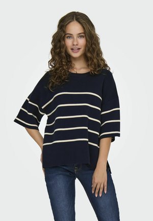 Pull oversized bleu marine avec des rayures horizontales blanches, col rond et manches courtes, associé à un jean bleu foncé. Texture en maille douce.