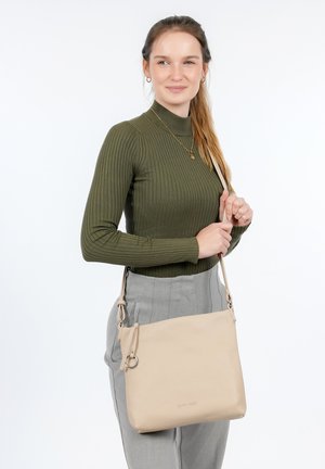 Borsa a tracolla in pelle color beige chiaro con finitura testurizzata, dotata di una tracolla regolabile e un logo frontale. La donna indossa un top rigato verde oliva e pantaloni grigi.