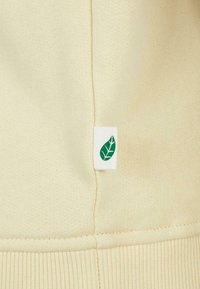 Tissu beige avec une couture verticale et une petite étiquette blanche avec un symbole de feuille verte sur le côté près de l'ourlet côtelé.