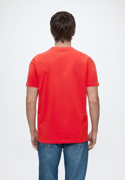 Homme portant un t-shirt rouge uni à manches courtes et un jean bleu, debout de dos sur un fond blanc uni.