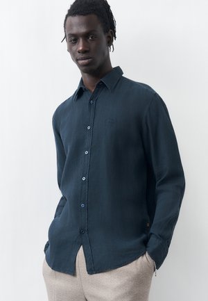 Νεαρός άνδρας με σκούρα πλεγμένα μαλλιά, που φοράει μπλε σκούρο πουκάμισο με κουμπιά και ανοιχτό μπεζ παντελόνι, στέκεται με τα χέρια στις τσέπες μπροστά σε λευκό φόντο.