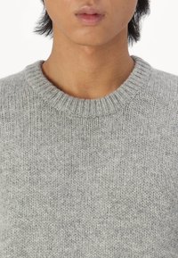 Pelēks adīts džemperis ar apaļu kakla izgriezumu. Audums izskatās mīksts un teksturēts, ar smalku rakstu un šķautņveida detaļām ap kaklu.