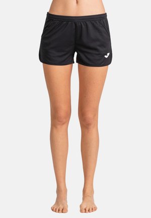 Piernas y pies desnudos de una persona que lleva pantalones cortos negros con cintura elástica y un pequeño logo blanco en el lado izquierdo, sobre un fondo liso.