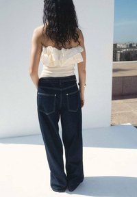 Jeans in denim scuro a gamba larga con cuciture visibili, due tasche posteriori e vita alta. Abbinati a un top in crema con maniche a sbuffo off-shoulder.