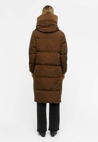 Cappotto lungo imbottito marrone con colletto alto, cuciture dettagliate e design trapuntato. Abbinato a pantaloni larghi scuri.
