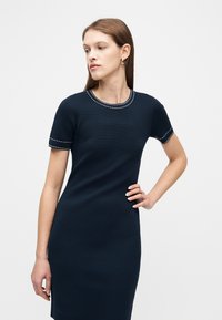 Γυναίκα με ίσια καστανά μαλλιά φοράει εφαρμοστό ναυτικό μπλε φόρεμα με λευκές ραφές στο γιακά και στα μανίκια, ποζάροντας με το χέρι στη μέση.