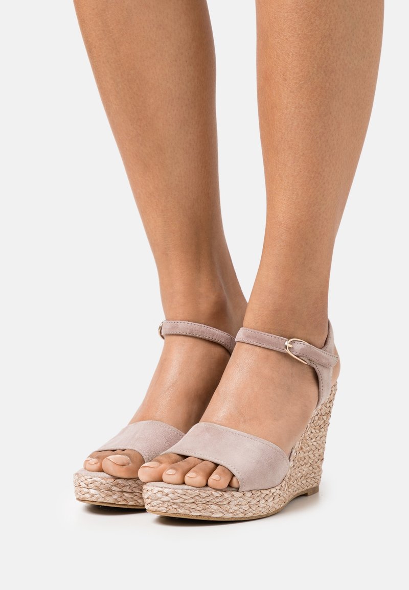 Pieds portant des sandales compensées en daim rose clair avec brides à la cheville et talons plateforme en jute tressée sur fond blanc.