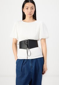 Svart kunstlær korsettbelte med øsken og snøring foran, brukt over en strukturert hvit topp, sammen med blå høytlivsjeans.