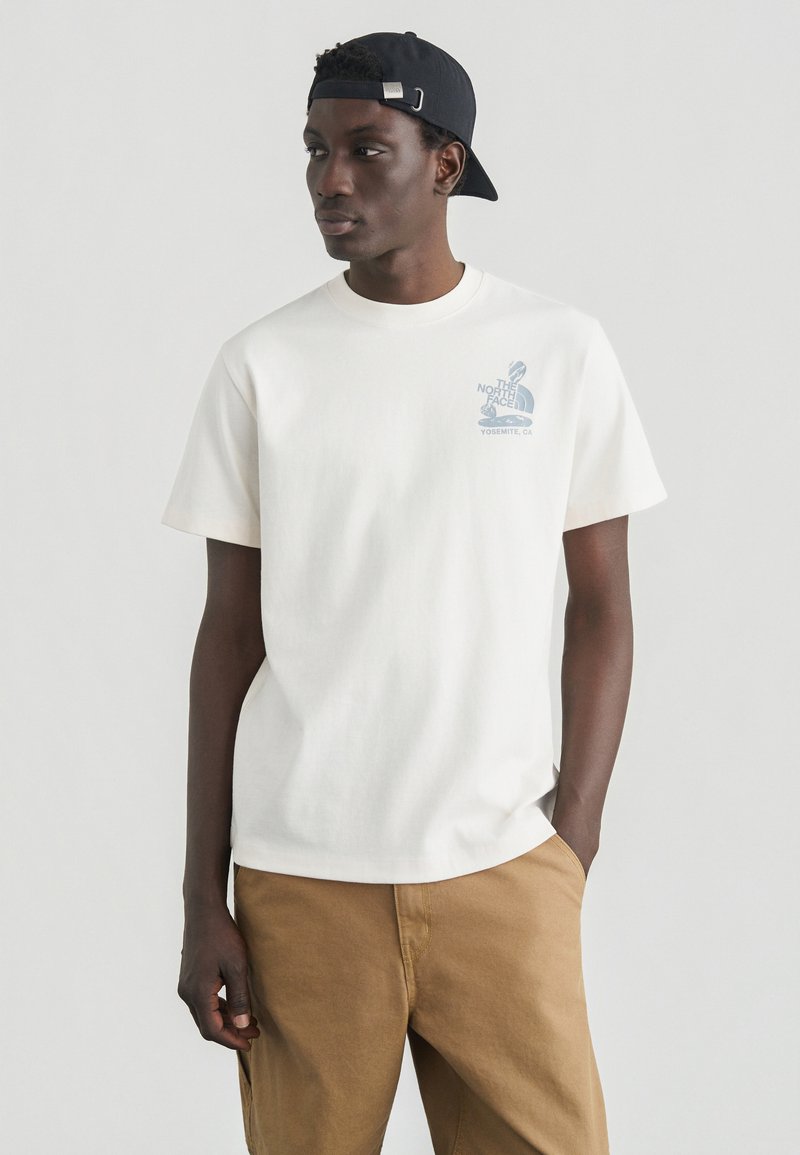 Λευκό βαμβακερό t-shirt με στρογγυλή λαιμόκοψη, κοντά μανίκια, που έχει μπλε γραφικό λογότυπο στο αριστερό στήθος. Στυλιζαρισμένο με χακί παντελόνια.