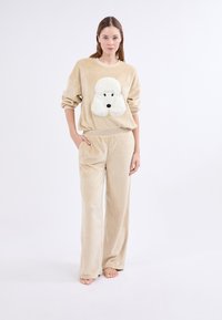 Ensemble de loungewear en polaire beige avec un graphique en peluche d'un caniche blanc sur le sweatshirt. Le pantalon a une coupe décontractée avec des poches latérales.