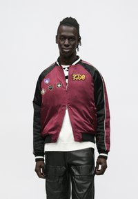 Burgundi värvi pommitaja jakk mustade varrukatega, millel on erinevad tikitud plaastrid ja soonikkoes ääris. Kantud valge pluusi ja mustade pükste peal.