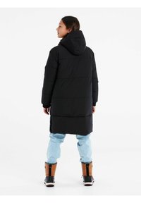 Giacca a vento nera con cappuccio e design lungo, caratterizzata da sezioni trapuntate. Indossata sopra jeans azzurri chiari e stivali invernali marroni.