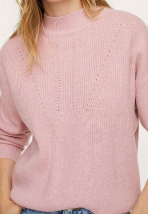 Femme portant un pull en maille rose clair avec de petits trous décoratifs en motif chevron et des manches retroussées.