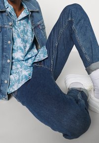 Chaqueta de mezclilla, camisa azul claro con estampado de palmeras, jeans azul oscuro, zapatillas blancas con detalles en gris, usadas con los puños remangados y costuras visibles.