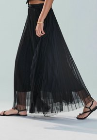 Falda negra plisada hecha de un tejido ligero y transparente. Llevada con sandalias negras planas que presentan un hardware minimalista. Diseño fluido hasta los tobillos.