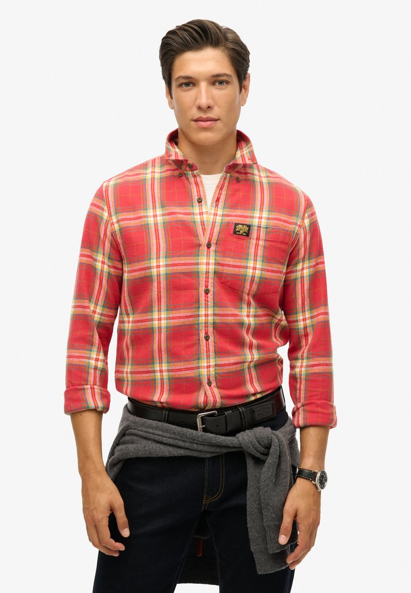 Rood geruit overhemd met gele, groene en blauwe accenten. Lange mouwen, met knoopsluiting en een logopatch. Donkerblauwe jeans en een grijze trui om de taille geknoopt.