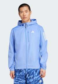 Veste zipée bleu clair en matériau léger, avec une capuche, des poignets élastiques et trois rayures blanches sur la manche gauche.