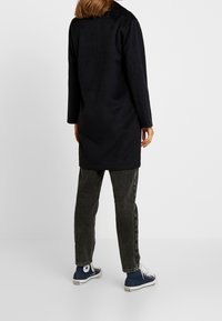 Zwarte wollen jas met lange mouwen, rechte snit en een gladde textuur, gecombineerd met grijze jeans en blauwe sneakers.