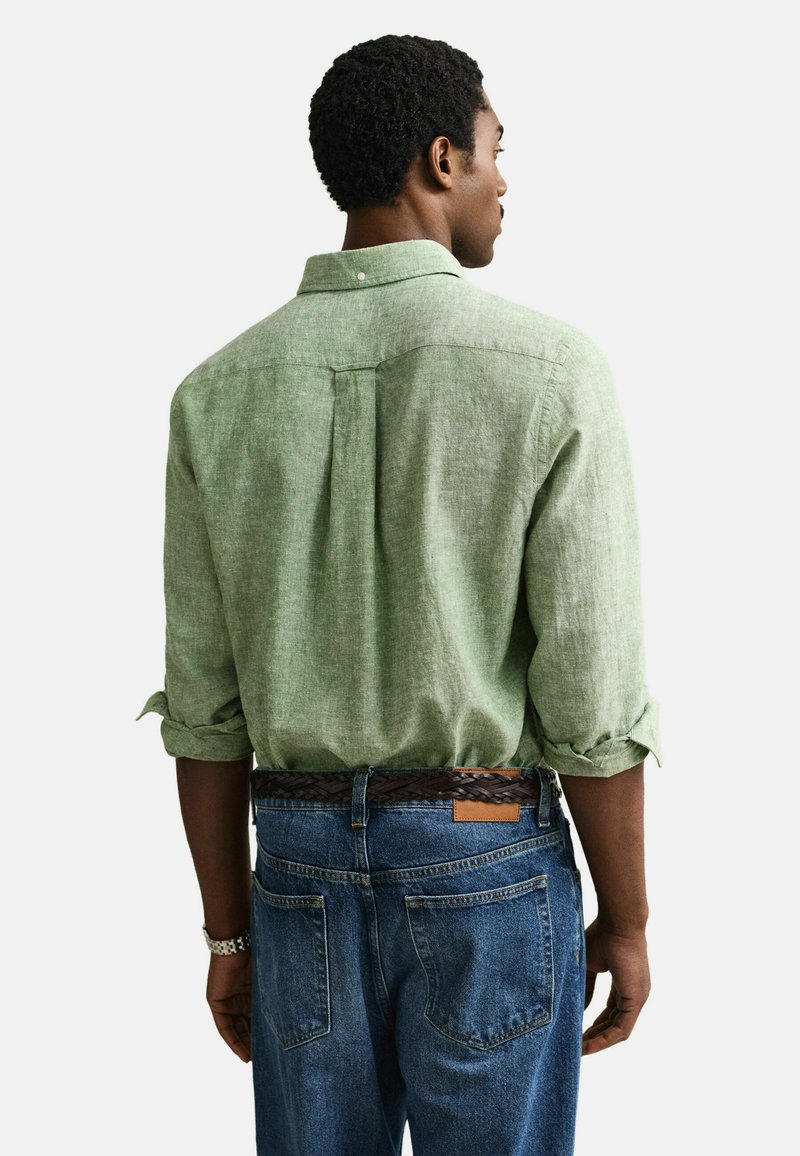 Hombre de espaldas que lleva una camisa de manga larga verde claro con los puños remangados y vaqueros azules con un cinturón trenzado marrón.