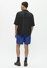 Cămașă neagră cu mânecă scurtă, cu o croială lejeră și o margine dreaptă, asortată cu pantaloni scurți albaștri. Ținuta include elemente de design subtile, fără modele vizibile.