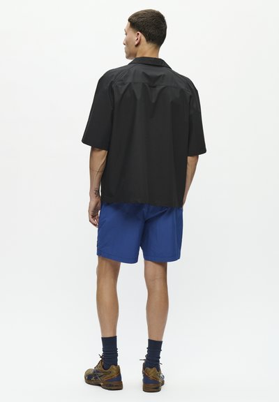 Black short-sleeve shirt with a relaxed fit and a straight hem, paired with blue shorts. The outfit features subtle design elements with no visible patterns.