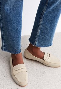 Mocasines de lona beige con un acabado texturizado y un sutil acento de costura, combinados con jeans de mezclilla azul deshilachados.