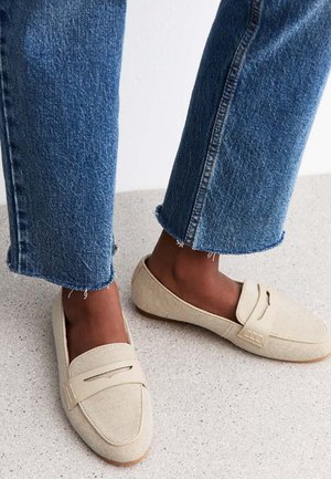 Mocassini in tela beige con una finitura texturizzata e un sottile dettaglio cucito, abbinati a jeans in denim blu sfrangiati.