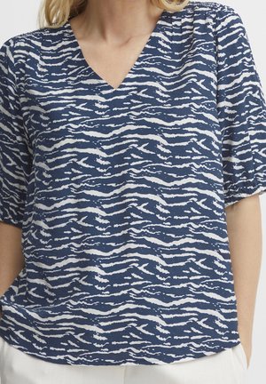 Kobieta w granatowo-białej bluzce z abstrakcyjnym, paskowanym wzorem, dekoltem w serek i krótkimi bufkowatymi rękawami, w połączeniu z jasnymi spodniami.