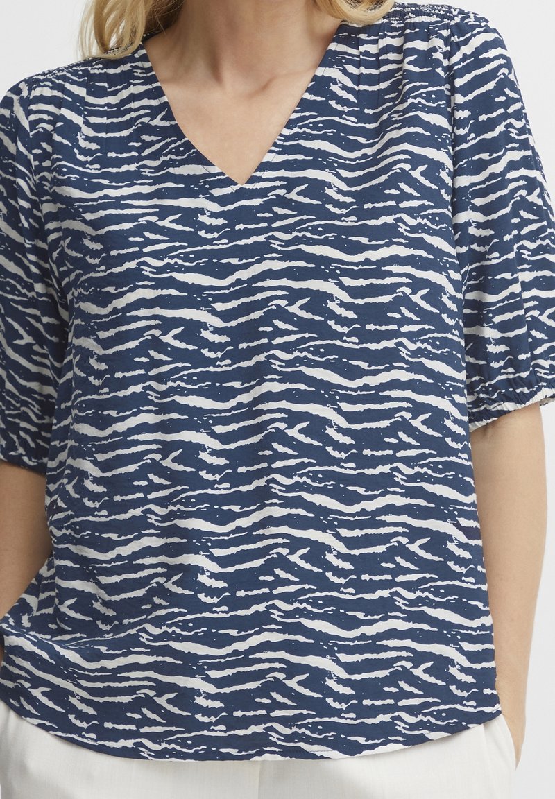 Femme portant un chemisier à rayures abstraites bleu marine et blanches avec un col en V et des manches courtes bouffantes, associé à un pantalon de couleur claire.