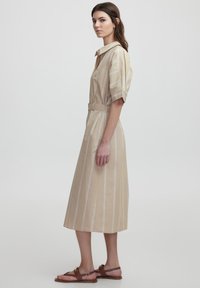 Abito camicia beige a righe con maniche corte, colletto, vita con cintura e silhouette ad A, abbinato a sandali piatti marroni.