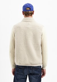 Man in een crème fleece vest over een langarmshirt, met een blauwe honkbalpet en donkerblauwe jeans, staand met zijn rug naar de camera tegen een witte achtergrond.