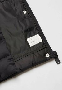 Schwarze Pufferjacke mit glattem Nylonfutter, einem Etikett für Name und Telefonnummer sowie einem Druckknopfdetail an der Seite.