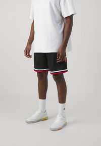 Pantaloni corti da basket in rete neri con rifiniture rosse e bianche; abbinati a una maglietta bianca larga e sneaker bianchi con dettagli neon.