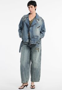 Model draagt een losse spijkerjack en baggy jeans, gecombineerd met zwarte sandalen, terwijl hij tegen een effen witte achtergrond staat en een casual stijl uitstraalt.