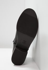 Chaussure noire avec une tige en cuir lisse et une semelle en caoutchouc texturée. Elle présente un petit talon et une étiquette de marque embossée sur le côté.