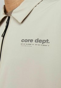 Giacca a zip beige chiaro con colletto a punta, con la scritta "core dept." e coordinate in grigio. Tessuto liscio con finitura opaca.