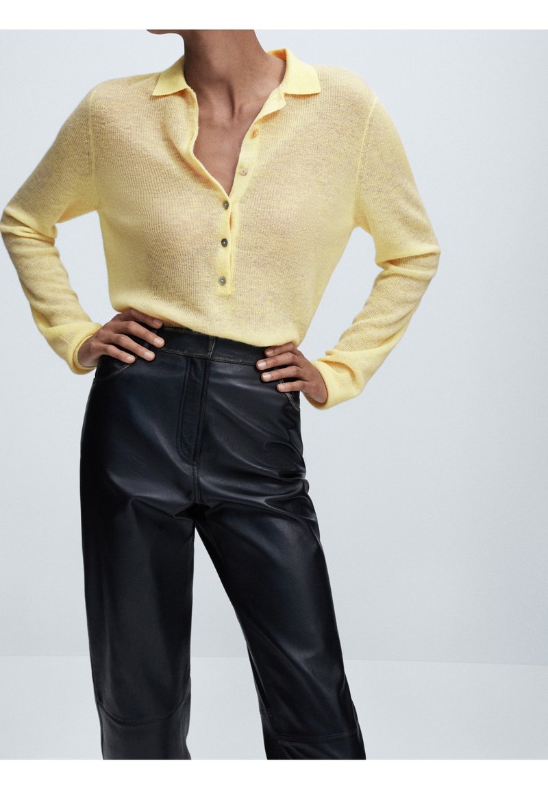 Femme portant un pull jaune boutonné, rentré dans un pantalon en cuir noir taille haute, debout les mains sur les hanches.