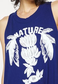 Femme portant un haut sans manches bleu avec un imprimé botanique blanc et le mot "NATURE" au-dessus du motif.