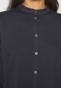 Gros plan sur une chemise bleu marine foncé avec cinq boutons visibles et un col rond portée par une personne aux cheveux longs.