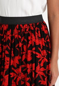 Jupe florale avec une base noire ornée de grandes fleurs rouges, design plissé et taille élastique noire. Texture de tissu lisse.