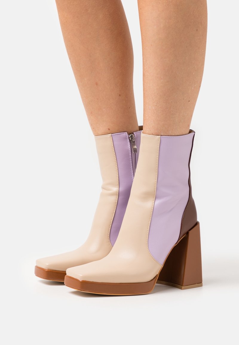 Bottes chelsea avec talons épais marron, panneaux avant beiges et panneaux latéraux lavande, portées sur des jambes nues sur un fond blanc.
