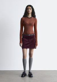 Rostfarbener Langarm-Top, burgunderfarbener Cordminirock, graue kniehohe Socken und schwarze Schuhe mit Quastendetail, vor einem weißen Hintergrund.